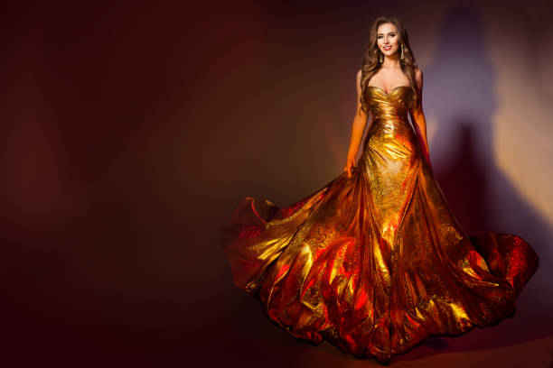 fashion model in long golden dress. smiling beauty woman in evening shining fluttering gold gown walking over dark brown background - sensuality walking women beautiful imagens e fotografias de stock