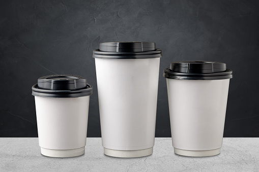 Three different sizes of white takeaway paper cups with black plastic lids isolated on gray background.