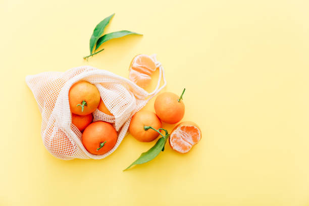 istock Fresh clementines with leaves in reusable string bag on yellow background, top view 1353196406