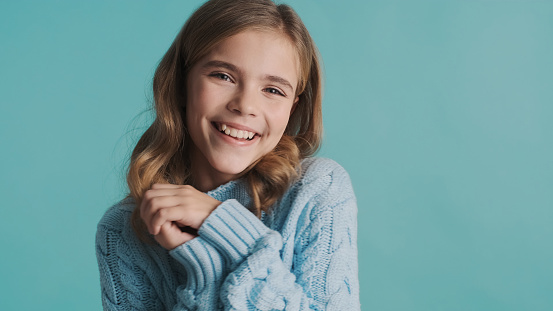 Beautiful cute blond teenage girl dressed in cozy sweater looking charming posing on camera over blue background. Face expression