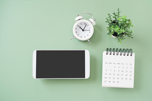 white smartphone, included clipping path on touchscreen,  white analog alarm clock, calendar, small green tree on green background for workspace concept