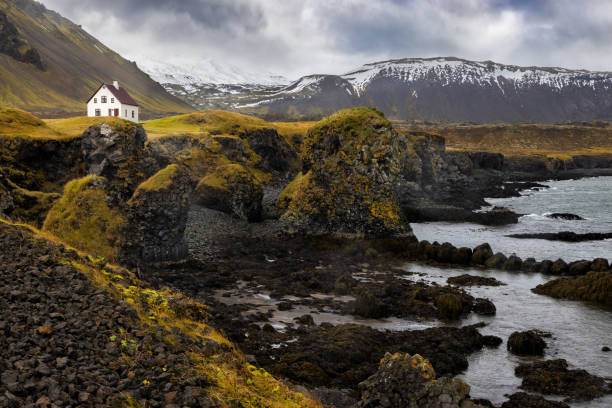 una vista de la escarpada costa de arnarstapi con una casa solitaria. colores otoñales con montañas cubiertas de nieve al fondo - snaefellsnes fotografías e imágenes de stock