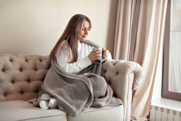 ソファの上のベージュのミニマルなリビングルームで自宅に座り、ニットのチェック柄の服を着た青いマグカップからお茶やコーヒーを飲む女性。居心地の良いヒュッゲ。暖かいセーターを� - blanket ストックフォトと画像