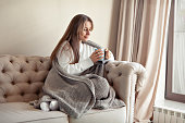 Woman sitting at home in beige minimalistic living room on sofa, drinking tea or coffee from blue mug, dressed in knitted plaid. Cozy hygge. Caucasian model with long hair wearing warm sweater