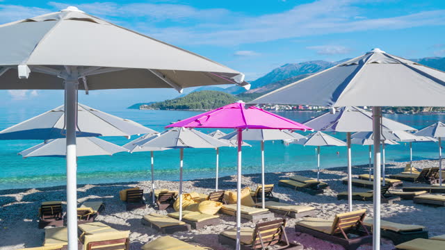 One pink beach umbrella among rest of grays. Umbrellas are gradually opening up. Girl comes and lies down on a sun lounger.