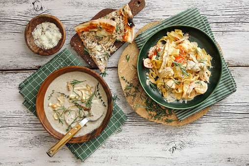 Italian fettuccine with prawns, salmon and herbs. Ham and mushroom pizza. Creamy vegan wild mushroom soup with herb and vegetable chips. Flat lay top-down composition on wooden background.