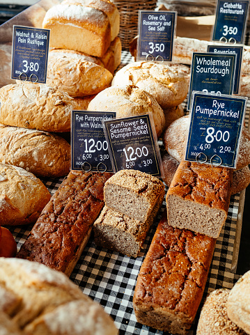 Color image depicting a large selection of artisan gourmet breads displayed on a bakery stall at a food market. The different breads include sourdough, rye and many others. Each item is labelled and priced. Room for copy space.