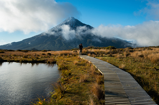 Hiker standing on the wooden boardwalk platform around the Pouakai tarn. Mt Taranaki in the background.