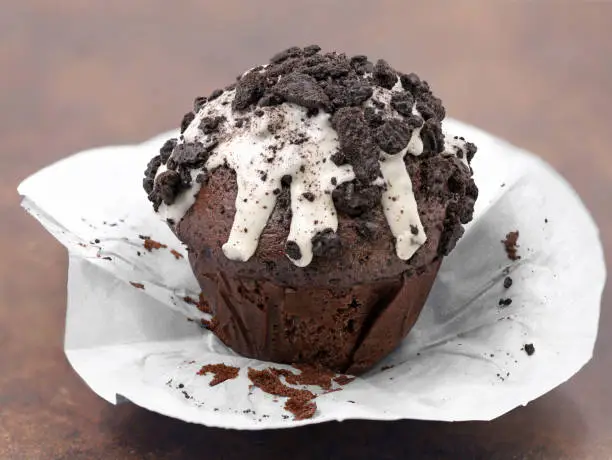 Photo of chocolate muffin with white glaze and dark cookie crumbs on brown background, unpacked cupcake