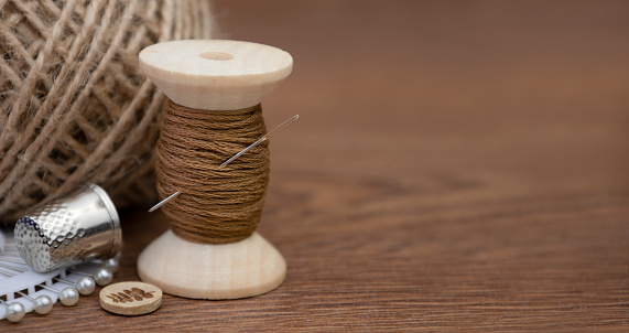 A spool of brown thread, a needle and a thimble on a wooden background.Accessories for sewing and needlework