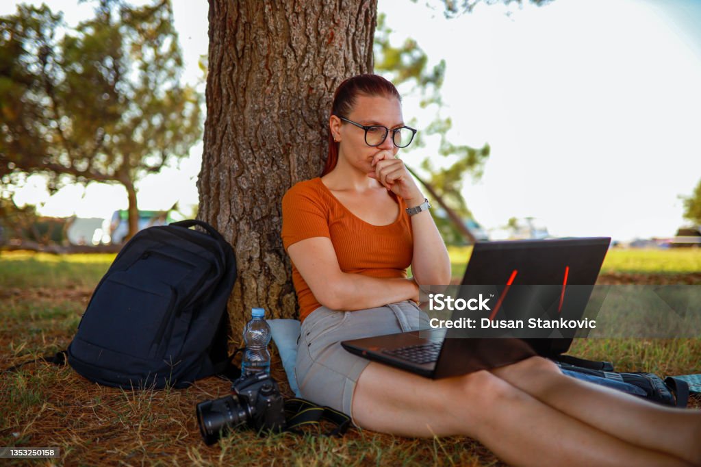 Focused woman, working on her laptop, while sitting on the grass int he public park Young woman, leaning on the tree in the public park while surfing the net on her laptop 25-29 Years Stock Photo