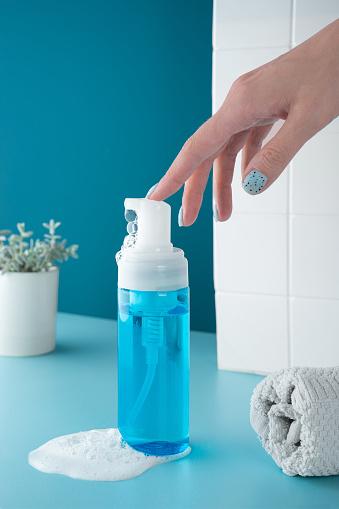 Beauty product mockup. Wellness packaging. Blue bottle, Cleaning cosmetic. Hand pumping cleansing foam in Plastic pump bottle. skincare routine. Bath background, glass with drops and a white towel