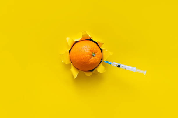 Small syringe are stuck in an orange fruit. Torn hole in yellow paper, copy space. The concept of vaccination, anti-cellulite injections and medical procedures. stock photo
