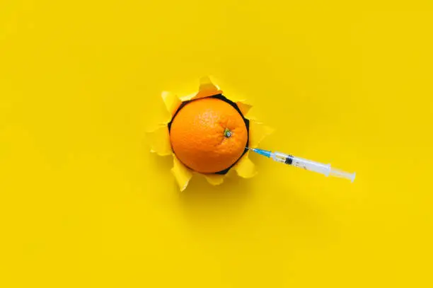 Small syringe are stuck in an orange fruit. Torn hole in yellow paper, copy space. The concept of vaccination, anti-cellulite injections and medical procedures.