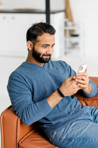 istock Serene Indian man holds smartphone sitting on the couch at home 1353257535