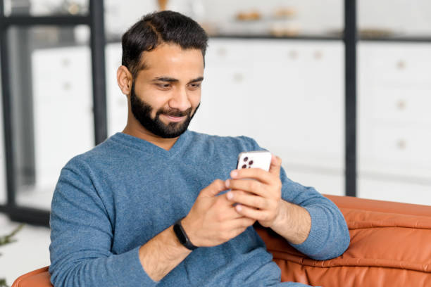 istock Focused bearded Indian man lying on the sofa at home and holding smartphone 1353257564