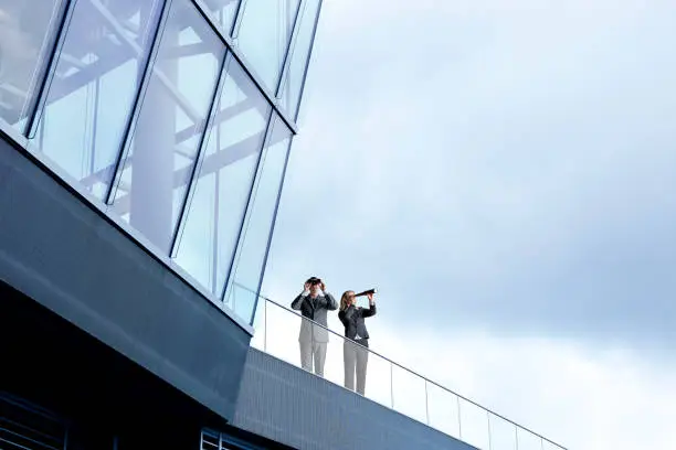 A businessman looks through a pair of binoculars while female colleague looks through a spyglass as they stand at the edge of a balcony of a modern office building.