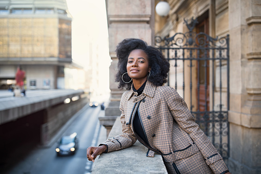 Beautiful African American young woman with afro and large hoop earrings in a stylish coat, smiling. City life concept, street portrait. Spring or fall time. Selective focus, copy space