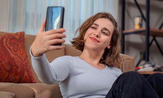 Pretty young female with big smile sitting at living room. beautiful young woman taking selfie while sitting on carpet at home. She having fun taking light cheerful selfie on blurred background. woman of average age of 20 years dressed casually is in the living room of her house sitting on the sofa taking a selfie with her cell phone.