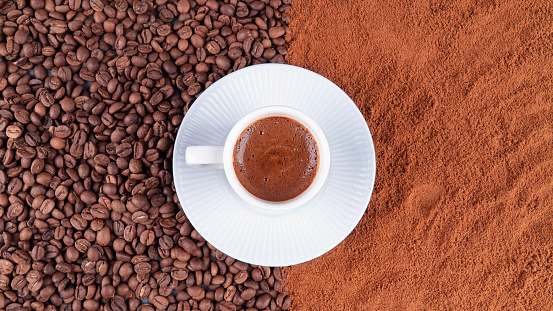 Turkish Coffee in roasted and ground coffee beans from top view. Traditional tasty refreshment turkish coffee from  turkish cuisine culture. White cup of turkish coffee on a white plate. Coffee beans all around. Top view. Traditional cup of Turkish coffee with foam on sharply separated ground and whole coffee beans background.