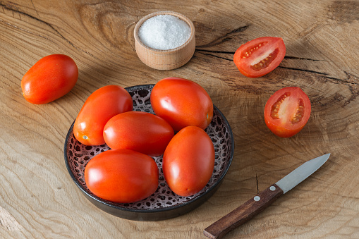 A lot of whole tomatoes in the shape of plums and one cut are in a plate and on a wooden board