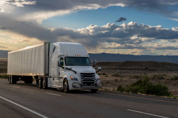 White Semi-Truck Speeding Down a Highway in the Utah Desert at Dusk under a dramatic sky stock photo
