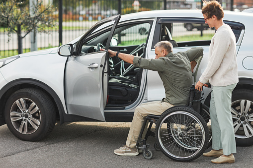 Portrait of woman helping man in wheelchair enter car in parking lot outdoors, copy space