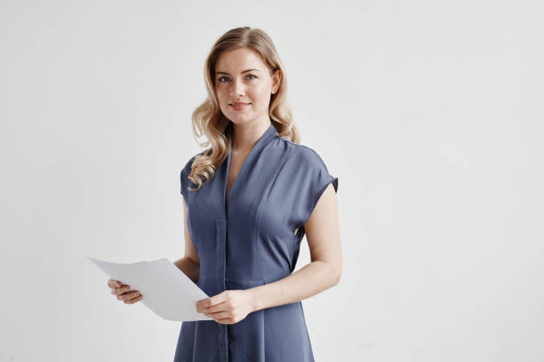Successful Woman Holding Documents on White Minimal waist up portrait of beautiful blonde businesswoman holding documents and looking at camera while standing by white wall, copy space paper stock stock pictures, royalty-free photos & images