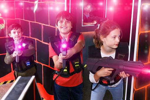 Positive teenage girl and boys with laser pistols playing laser tag on dark labyrinth