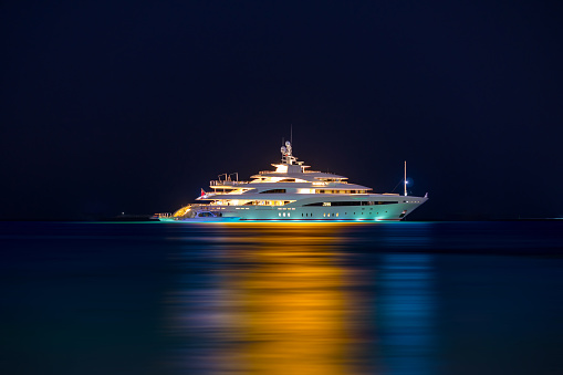 Night view to large illuminated white boat located over horizon, colorful lights coming from yacht reflect on the surface of the the Gulf sea. Shot at blue hour.