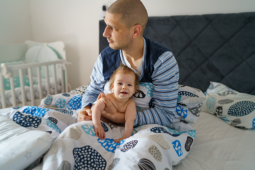 Baby and father adult caucasian man hold his six months old daughter girl on the bed wearing blue pajamas in the morning at home parenthood family domestic life bonding concept real people copy space
