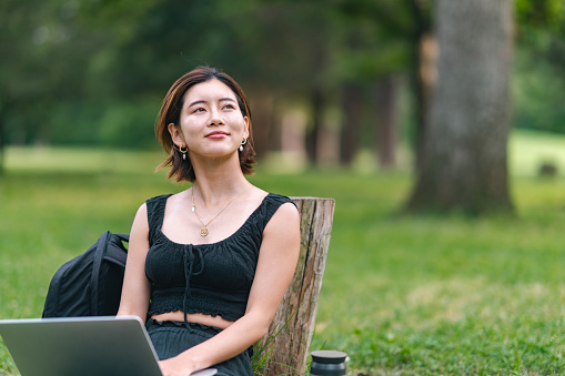 A young woman is relaxing in nature and using a laptop. A portrait of a young woman is looking away and contemplating while using a laptop in nature.