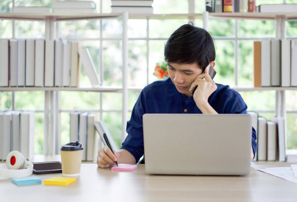 istock Young asian man take a note while listening on mobile phone. Work from home concept. 1353333915