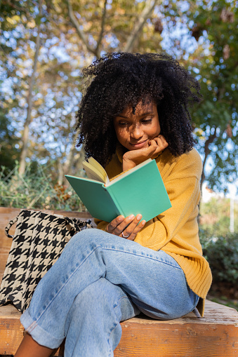Vertical portrait of young African American reading a book sitting on a park bench. Lifestyle concept.