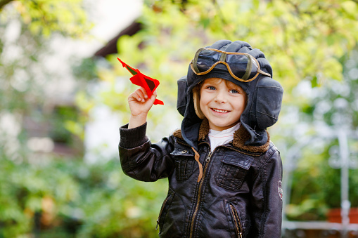Happy little kid boy in pilot helmet and uniform playing with red toy airplane. Smiling preschool child dreaming and having fun. Education, profession, dream concept.