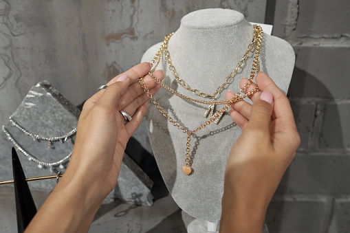 Hands of young female consumer holding gold necklace on mannequin while choosing jewelry in luxurious boutique