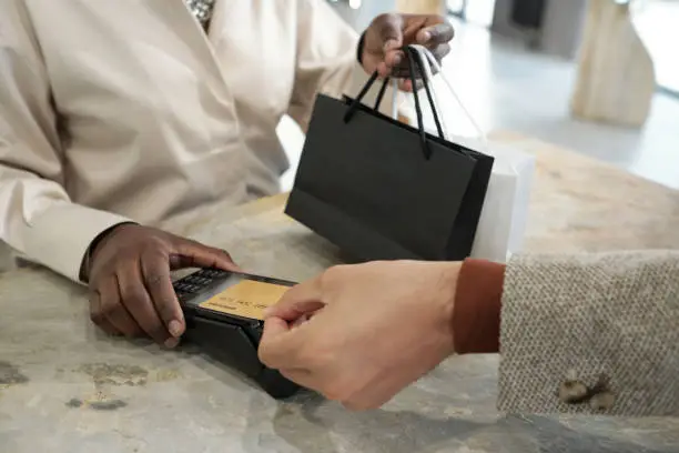 Hand of young man holding credit card by screen of payment terminal while buying gifts in jewelry boutique and paying for purchase