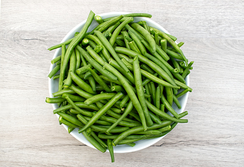 Raw green beans in a white bowl on wooden table.  View from above