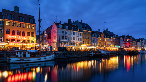 Copenhagen Nyhavn Nightlife Panorama in the famous illuminated Nyhaven District of Copenhagen in moody sunset twilight. Crowds of People walking along the channel to visit Restaurants and Bars in colorful traditional danish houses. Unrecognizable Crowd of People, several cityscape logo signs not in focus. Nyhavn, Copenhagen, Denmark, Nordic Countries, Europe