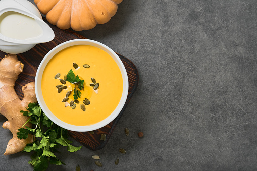 Pumpkin carrot vegetarian soup with parsley, olive oil and pumpkin seeds in a white bowl, top view. Layout on gray background with ingredients, copy space for menu