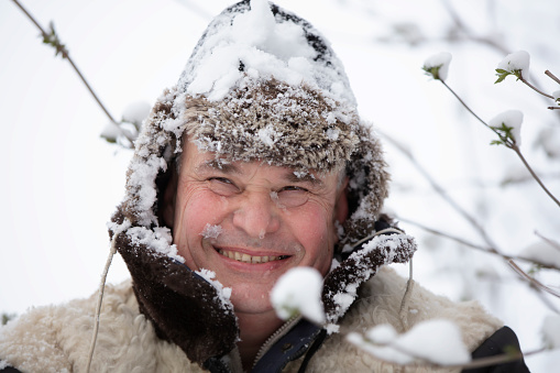 An elderly man in a winter hat covered with snow smiles and looks into the camera.