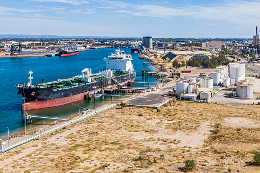 Aerial view of bulk tanker ship moored at petrochemical  terminal berth in river harbor, discharging fuel into white petrochemical storage tanks. In the background is an extensive industrial district, new residential development, with the coast and city suburbs in the far distance.  ID & logos edited.