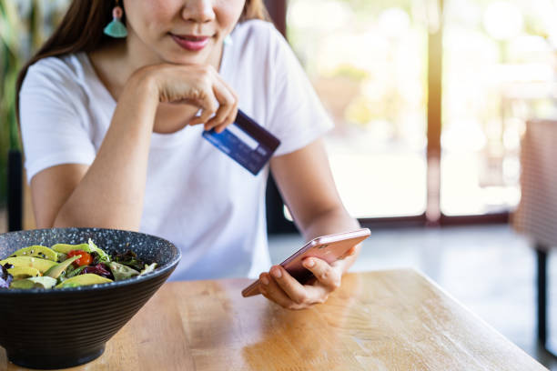 Young asian woman holding a credit card and using smart phone for make an online shopping payment at restaurant stock photo