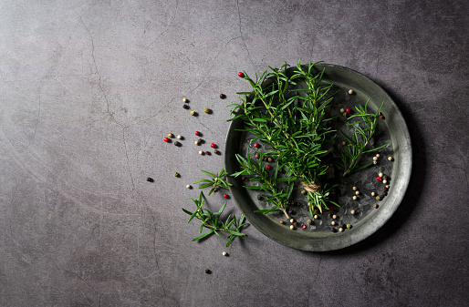 A bunch of fresh rosemary and pepper mix on an iron plate set against a dark background. View from directly above.