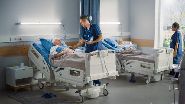 hospital ward: friendly male nurse talks reassuringly to elderly patient resting in bed. doctor or physician uses tablet computer, does checkup, old man fully recovering after successful surgery - bed stockfoto's en -beelden