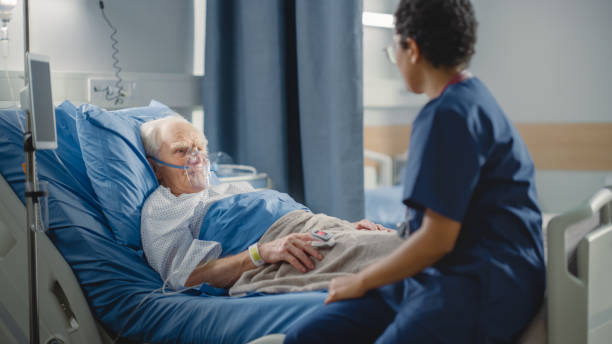 病院病棟:フレンドリーヘッドナースは、ベッドで休んでいる酸素マスクを身に着けている高齢患者に指の心拍数モニターまたはパルスオキシメータを接続します。看護師は、手術に成功した - hospital patient doctor bed ストックフォトと画像