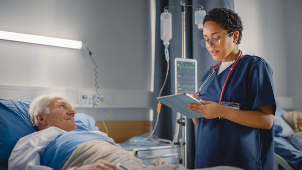 Hospital Ward: Friendly Head Nurse Talks with Elderly Patient Resting in Bed. Physician Uses Tablet Computer, Does Checkup, Ask Health Care Questions. Old Man Fully Recovering after Successful Surgery Hospital Ward: Friendly Head Nurse Talks with Elderly Patient Resting in Bed. Physician Uses Tablet Computer, Does Checkup, Ask Health Care Questions. Old Man Fully Recovering after Successful Surgery hospital patient bed nurse stock pictures, royalty-free photos & images