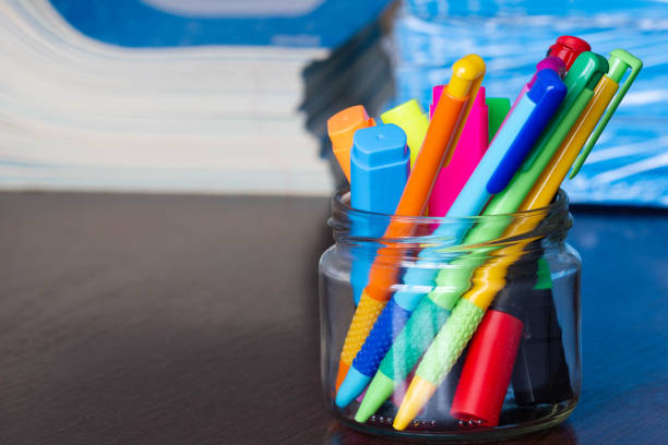 istock Multi-colored pens and markers in a stand on the background of Stack of Notebooks.  Pack of blue school notebooks on the table in a classroom. Stack of exercise books. Packing checkered math notebooks 1353358880