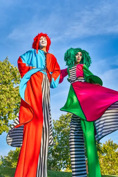 From below of funny show performers on stilts in colorful clown costumes and wigs standing together in green park and during festive event