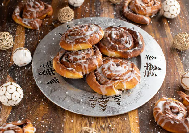 Fresh and homemade baked traditional cinnamon rolls with glaze. Served on a silver christmas plate with decoration on wooden and rustic table from above